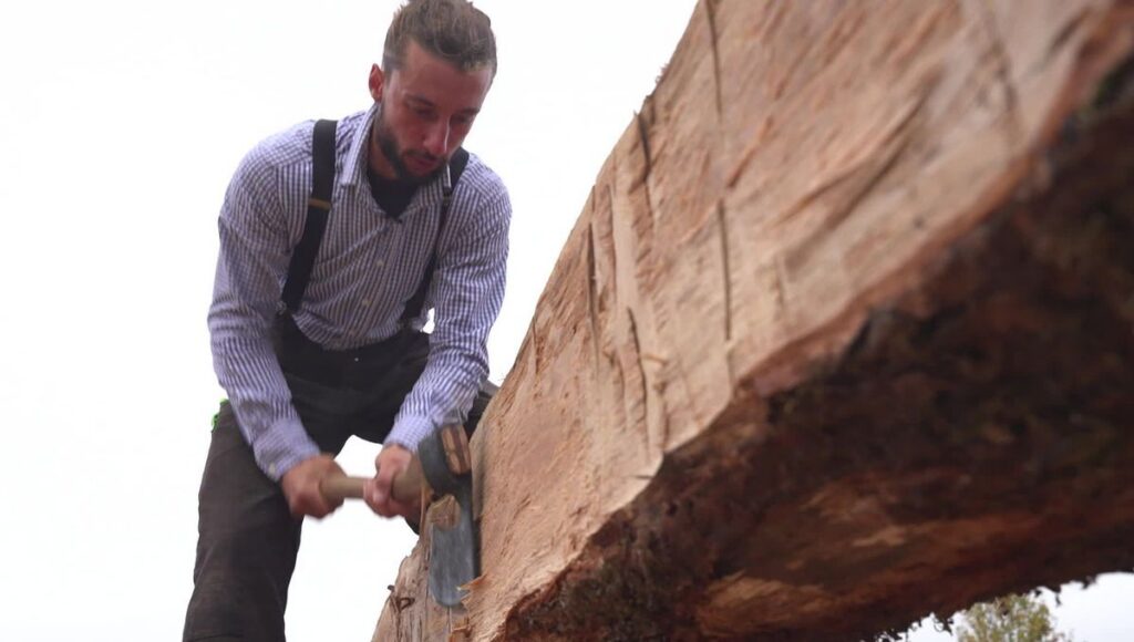 Charpentier travaillant à la hache une pièce de charpente en bois, geste hérité des bâtisseurs nordiques